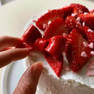 Extreme close-up of a hand placing a glossy red fondant bow on a white cake with pink hearts and strawberries. (Hello Kitty Birthday Party Ideas)